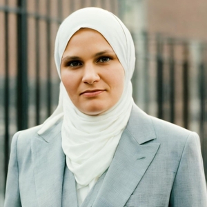 A woman in a white headscarf and light gray blazer faces the camera outdoors.  