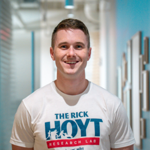 A young man with short brown hair is smiling confidently at the camera while standing in a bright hallway with blue walls and framed artwork. He is wearing a white t-shirt featuring “The Rick Hoyt Research Lab” logo in blue and red.