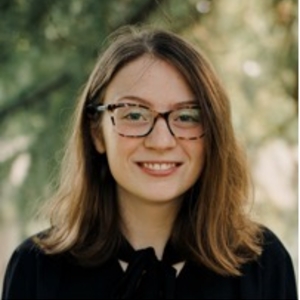 A young white person with medium-length brown hair and glasses smiles at the camera. She poses in a black shirt for a professional photograph in front of a leafy outdoor background. 