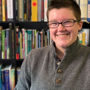 A young white person with short brown hair and black framed glasses is smiling directly at the camera. They are wearing a gray turtleneck sweater with three wooden-toned buttons down the front. They are against a backdrop of tall, black bookshelves tightly packed with books of different colors and sizes.