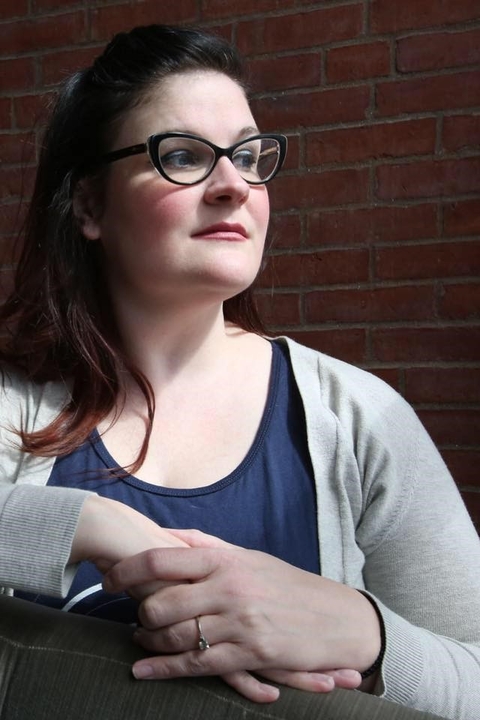 A person with long hair and glasses, wearing a blue shirt and a light gray cardigan sweater. 