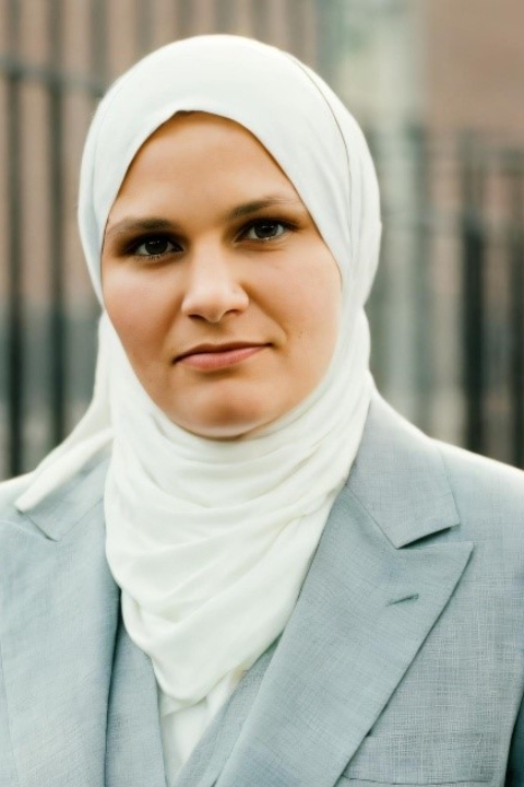 A woman in a white headscarf and light gray blazer faces the camera outdoors.  