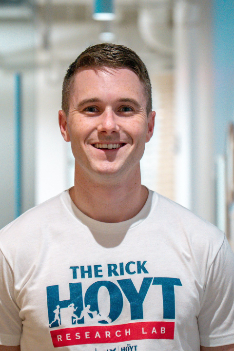 A young man with short brown hair is smiling confidently at the camera while standing in a bright hallway with blue walls and framed artwork. He is wearing a white t-shirt featuring “The Rick Hoyt Research Lab” logo in blue and red.
