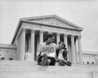 A black and white photograph of an African American woman sitting on the steps of the Supreme Court, looking affectionately at her daughter. The woman is holding a newspaper with the headline 'High Court Bans Segregation in Public Schools.'