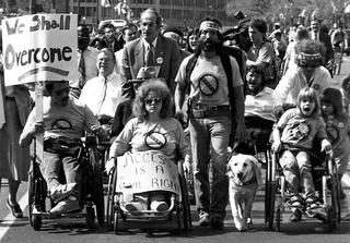 People engaging in a protest march for disability rights. Several people in wheelchairs are at the front of the line with signs such as We Shall Overcome and and Access is a Civil Right.